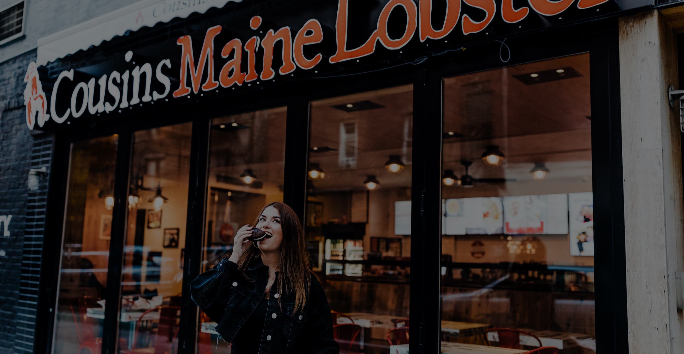 An image of a Cousins Maine Lobster storefront, with a woman outside enjoying a chocolate whoopie pie.